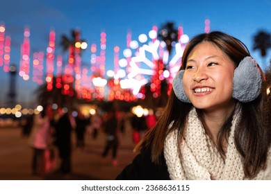 Happy young Asian woman wearing ear muffs and scarf on a cold winter night looking away. Copy space. - Powered by Shutterstock