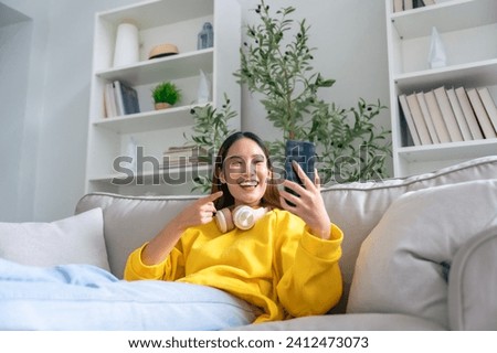 Happy young asian woman  wearing headset and relaxing on comfortable couch at home, texting messaging on smartphone, smiling girl use cellphone chatting, online shopping at home, video call