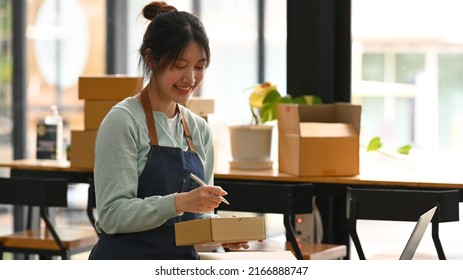 Happy Young Asian Woman Writing Address On Cardboard Box, Working For Online Selling A Home Office. Concept Of Online Selling, E-commerce