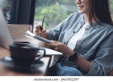 Happy young asian woman writing on notebook, working on laptop computer at coffee shop. Student studying online and taking note, surfing the internet, connecting wifi at cafe, business casual - Powered by Shutterstock