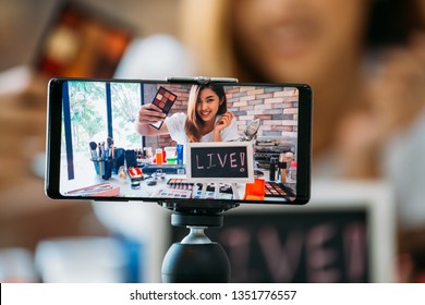 Happy young attractive Asian ethnic model filming vlog on mobile phone on tripod and showing eyeshadows at table with cosmetics