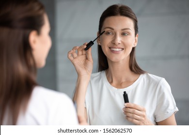 Happy Young Attractive Woman Holding Brush Applying Mascara On Eyelashes Getting Ready In Morning, Smiling Beautiful Lady Looking In Mirror Doing Make Up Routine Put Cosmetics On Face In Bathroom