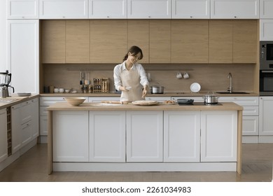 Happy young baker girl preparing pizza  pie  dropping grated cheese rolled dough  baking in home kitchen modern interior  enjoying culinary hobby  cooking activity  smiling