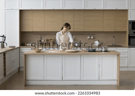 Happy young baker girl preparing pizza, pie, dropping grated cheese on rolled dough, baking in home kitchen modern interior, enjoying culinary hobby, cooking activity, smiling