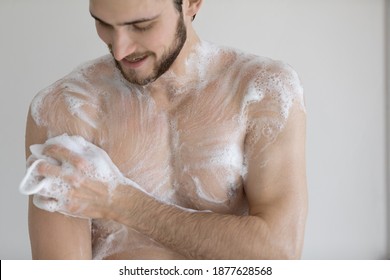 Happy young bare man cleaning body with foamy moisturizing gel, taking shower in bathroom. Smiling millennial guy enjoying morning or evening hygienic skincare routine, massaging body with washcloth.