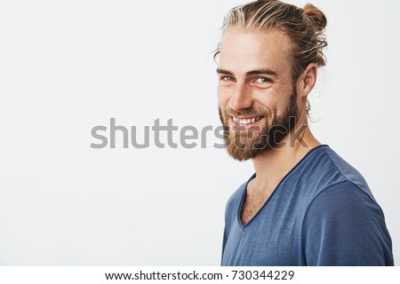 Happy young bearded guy with fashionable hairstyle and beard looking at camera, brightfully smiling with teeth, being happy about day off on work.