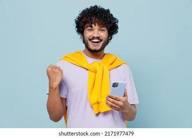 Happy Young Bearded Indian Man 20s Wears White T-shirt Hold In Hand Use Mobile Cell Phone Doing Winner Gesture Celebrate Clenching Fists Isolated On Plain Pastel Light Blue Background Studio Portrait