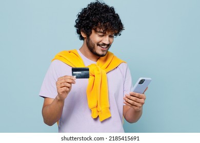 Happy Young Bearded Indian Man 20s Wears White T-shirt Using Mobile Cell Phone Hold Credit Bank Card Doing Online Shopping Order Delivery Isolated On Plain Pastel Light Blue Background Studio Portrait