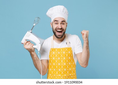 Happy young bearded male chef or cook baker man in apron white t-shirt toque chefs hat isolated on blue background. Cooking food concept. Mock up copy space. Hold kitchen mixer, doing winner gesture