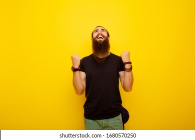 Happy Young Bearded Man In Casual Black Tee Shirt Posing Isolated On Yellow Background. Studio Portrait. People Lifestyle Concept. Copy Space. Clenching Fists Like Winner, Rising Hands. 