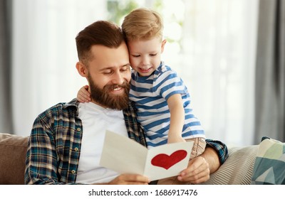 Happy Young Bearded Man In Casual Wear Reading Greeting Card While Sitting With Little Son On Sofa At Home In Fathers Day
