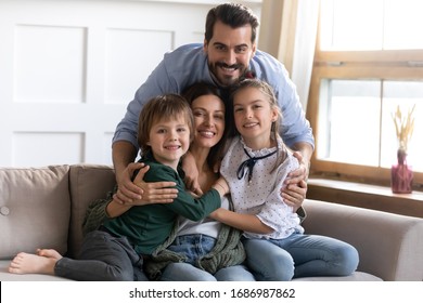 Happy Young Bearded Man Cuddling Sitting On Cozy Couch Beautiful Wife With Small Daughter And Son. Smiling Loving Full Family Posing For Photo In Living Room At Home, Good Relationship Concept.