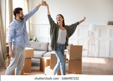 Happy young bearded man holding hand, twisting joyful wife in new living room near carton boxes. Excited married couple celebrating moving day in apartment house, dancing to energetic music.