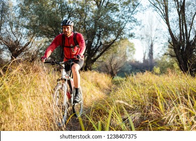 Happy Young Bearded Man In Red Long Sleeve Cycling Jersey Riding Mountain Bike Along A Path Through Tall Grass. Low Angle Front View.