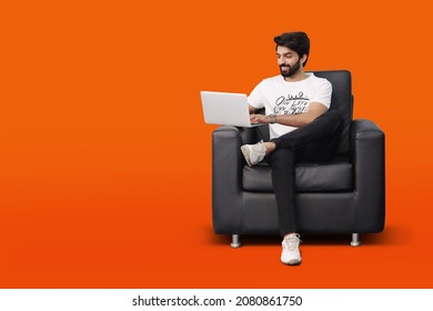 Happy young bearded man sitting on black sofa couch chair using laptop on orange background - Pakistani Indian South Asian - Shutterstock ID 2080861750