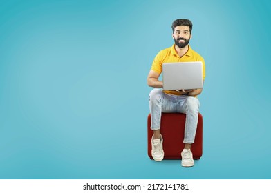 Happy And Young Bearded Man Sitting On A Block Stool Using Laptop On Blue Background - With Copy Space -  Pakistani Indian South Asian Middle Eastern