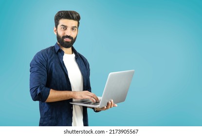 Happy Young Bearded Man Standing And Using A Laptop On Blue Background - Indian, Pakistani, South Asian, Middle Eastern