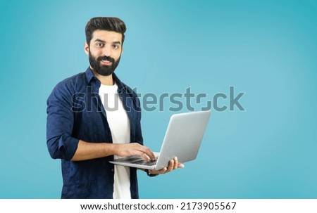 Happy young bearded man standing and using a laptop on Blue background - Indian, Pakistani, South Asian, Middle Eastern