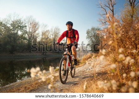 Happy young bearded man wearing red long sleeve cycling jersey and a backpack enjoying late afternoon mountain bike ride through the forest by river on a clear autumn day