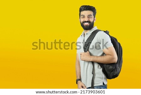 Happy young bearded student standing with a backpack on his shoulder on yellow background - Indian, Pakistani, South Asian, Middle Eastern
