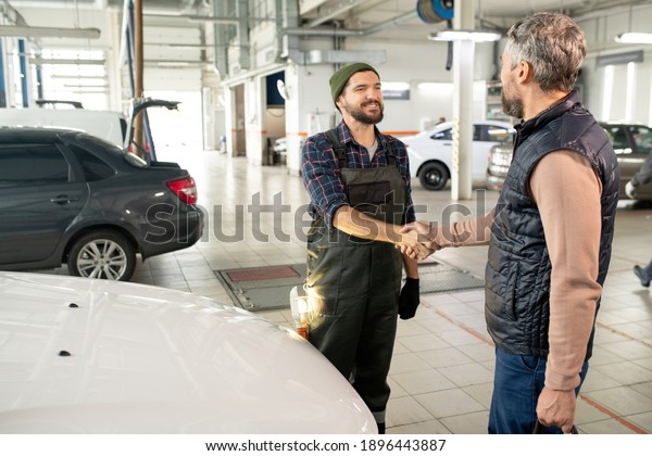 Happy young bearded technician of car service\
shaking hand of male client against front part of white car after\
checking it up and\
repairing