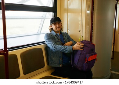 Happy Young Bearded Traveler Man In Baseball Hat And Jeans Jacket With Purple Backpack Sitting Inside Metro Train In European Country. Tourism, Traveling, Underground Or Lifestyle Concept.