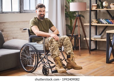 Happy young bearded veteran sitting in a wheelchair at his home. Disability and full life after military service.