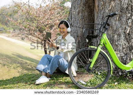 Happy young beautiful asian woman in casual clothes and headphone sitting beside big tree and bicycle and using smartphone to take selfie or having video call. Relaxation, nature, technology concept.