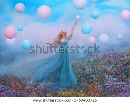 happy young beautiful blonde woman raised hand. Fantasy princess jumping touches pink ball air balloon. long blue tulle dress fluttering fly in wind. white clouds, fog, smoke colorful flowers meadow