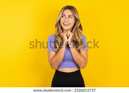 Happy Young beautiful blonde woman wearing sportswear over yellow studio background anticipates something awesome happen, looks happily aside, keeps hands together near face, has glad expression.