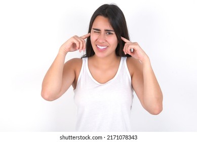 Happy Young Beautiful Brunette Woman Wearing White Top Over White Wall Ignores Loud Music And Plugs Ears With Fingers Asks To Turn Off Sound