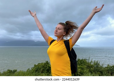 Happy young beautiful carefree woman traveler is hiking, travelling, enjoying freedom, walking on top of mountain with sea, beach view, breathing deep deeply fresh sea, ocean air, rising hands up