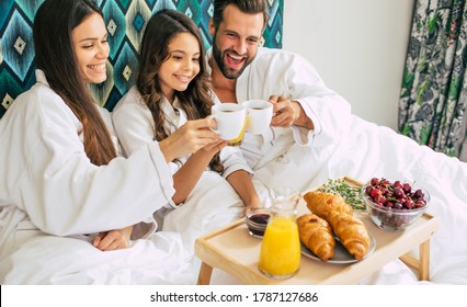 Happy Young Beautiful Family In Bathrobes Are Taking Morning Breakfast In A Luxury Hotel Room. Service In The Room. Vacation And Trip