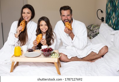 Happy Young Beautiful Family In Bathrobes Are Taking Morning Breakfast In A Luxury Hotel Room. Service In The Room. Vacation And Trip