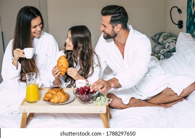 Happy Young Beautiful Family In Bathrobes Are Taking Morning Breakfast In A Luxury Hotel Room. Service In The Room. Vacation And Trip