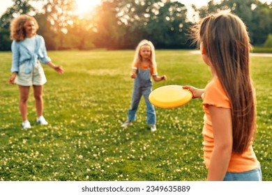 Happy young beautiful mom playing frisbee with her two beloved daughters in the meadow in the park, having fun together on a warm sunny weekend.