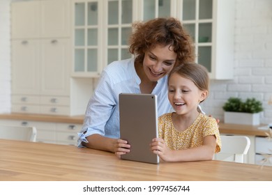 Happy young beautiful mother watching funny cartoons on digital computer tablet with joyful laughing little cute kid daughter playing online games, using applications together in modern kitchen. - Powered by Shutterstock