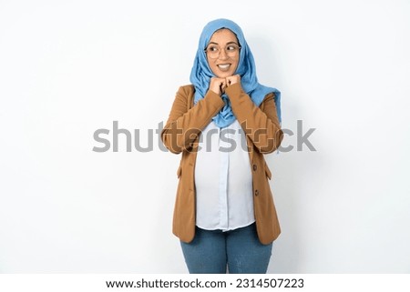 Happy Young beautiful pregnant muslim woman wearing hijab over white studio background anticipates something awesome happen, looks happily aside, keeps hands together near face, has glad expression.
