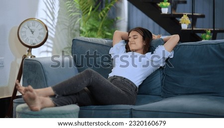 Happy young beautiful smiling woman sitting resting  on sofa raising arms stretching back muscles enjoying peaceful weekend at home. Indian female leaning on couch with closed eyes  hands behind head