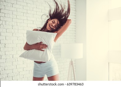 Happy young beautiful woman jumping on the bed at home, hugging pillow.