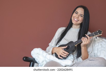 Happy Young Beautiful Woman Musician Playing On Ukulele Writing Music Sitting On Comfortable Couch At Home. Smiling Female Enjoying Hobby With String Musical Instrument Spending Time At Cozy Interior