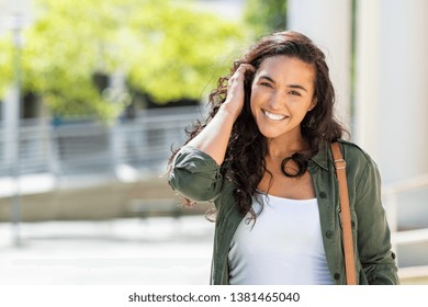 Happy young beautiful woman walking on the street. Portrait of cheerful university student looking at camera while adjusting curly hair with copy space. Latin stylish girl smiling while standing. - Shutterstock ID 1381465040