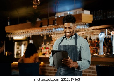 Happy young biracial man in apron using digital tablet while working as waiter in restaurant - Powered by Shutterstock