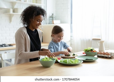 Happy Young Biracial Mother And Little Caucasian Daughter Chop Vegetables Prepare Healthy Salad At Home Kitchen, Smiling African American Mom And Small Girl Child Cooking Making Breakfast Together