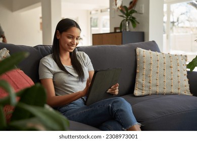 Happy young biracial woman in casual clothing using digital tablet on sofa in living room - Powered by Shutterstock