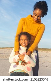 Happy Young Biracial Woman Embracing Daughter From Behind While Standing At Beach Against Sky. Family, Lifestyle And Weekend.