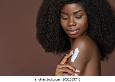 Happy young black African woman model with curly afro hair applying sunscreen lotion on shoulder isolated on brown background. Body skin care, sunblock and moisturizing bodycare treatment concept.