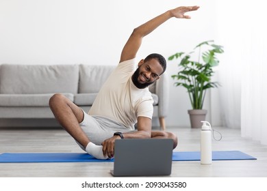 Happy Young Black Bearded Athlete Man In White Clothes Do Stretching Or Exercises For Hands On Mat, Looks At Pc In Living Room Interior. Online Lesson And Workout At Home Gym With Coach Due Covid-19