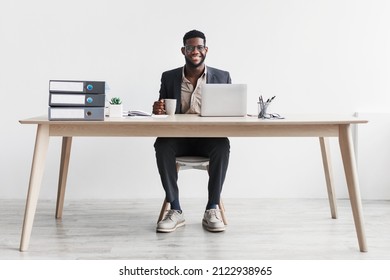 Happy young black businessman with coffee working online on laptop, sitting at desk and smiling at camera against white studio wall. Cheerful African American CEO enjoying remote job - Powered by Shutterstock