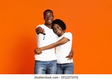 Happy Young Black Couple With New Car Keys Hugging And Smiling Over Orange Studio Background, Copy Space. Cheerful African American Man And Woman Embracing And Showing Key From Auto